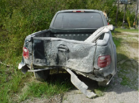 Dented rear ended black ford pick up truck sitting on the side of the road in the grass.