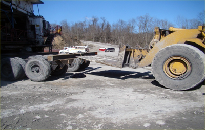 a miner transport dolly parked in front of a front-end loader. The arm of the loader is positioned within 2 feet of the back frame of the dolly.
