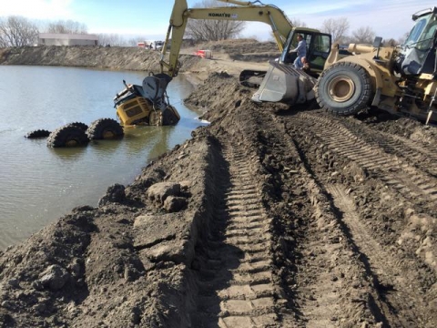 On a muddy shoreline, a crane positions its arm on a haul truck which is partially sunk into the water.